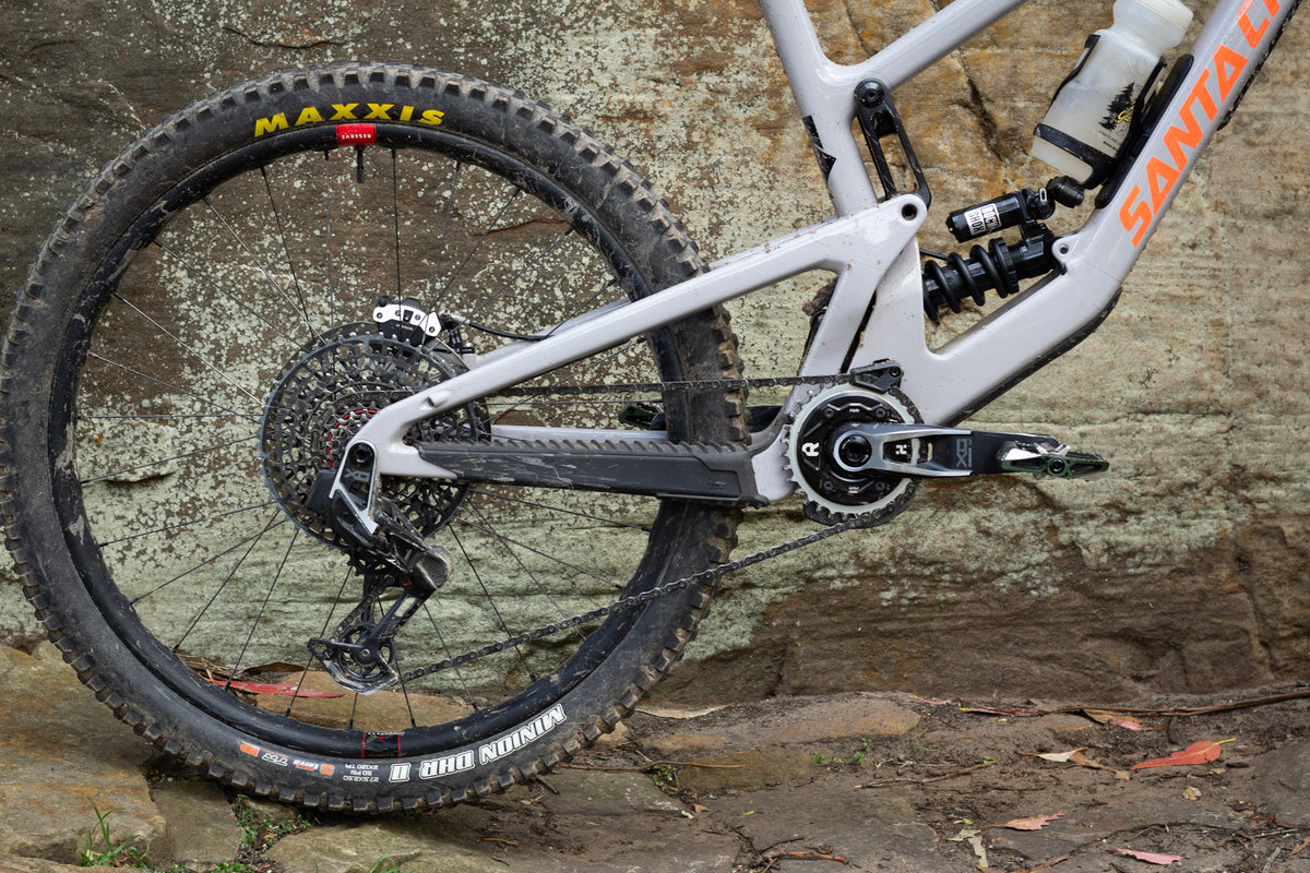 Close-up of a white Santa Cruz full-suspension mountain bike rear wheel and frame with a Rimpact Chain Damper, parked against a textured stone wall.