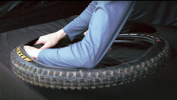 A person in a blue long-sleeve shirt is shown inside a rugged black mountain bike tire, demonstrating the interior fit or fitment of the tire. The tire sidewall features yellow and black Maxxis branding.