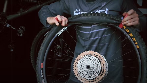 A person wearing a dark gray long-sleeve shirt holds a mountain bike wheel with a black Maxxis tire and a copper-colored cassette in a dark workshop setting.