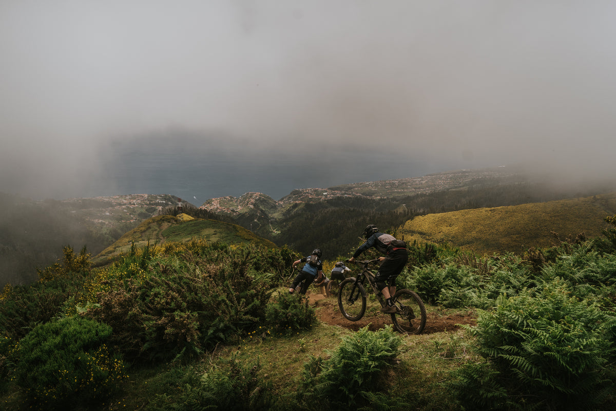 Two mountain bikers on a narrow dirt trail surrounded by green ferns and grassy hills, with misty mountains in the background under an overcast sky.