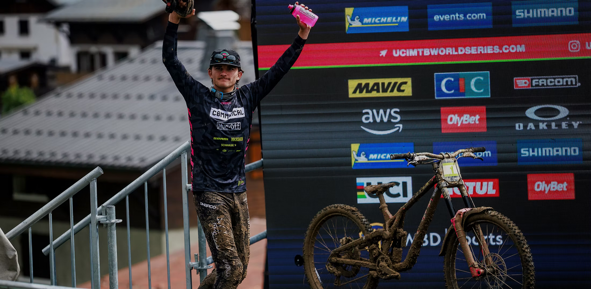 Max Alran on a podium with both arms raised, holding a pink water bottle, with a branded backdrop behind them and a muddy bike in the foreground.