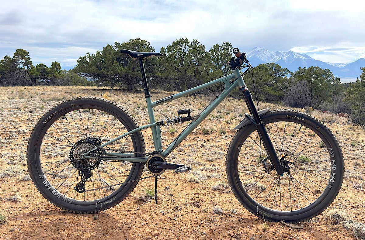 A full-suspension mountain bike with a matte sage green frame and black components, photographed outdoors on a dirt trail with desert shrubs, trees, and distant mountains under a cloudy sky.