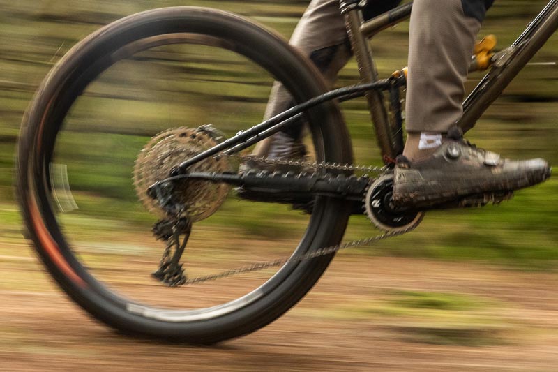 Close-up action shot of a mountain bike wheel and drivetrain in motion on a dirt trail, with a rider's legs and shoes visible against a blurred green outdoor background.