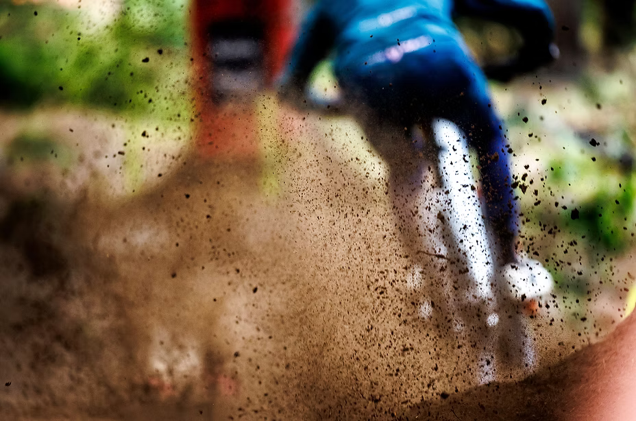 A dynamic, action-filled shot of a cyclist riding through deep mud and splashing dirt, viewed from behind. The rider is wearing a blue top and dark pants, navigating a muddy path with a dramatic spray of brown mud and black splatters in the foreground.