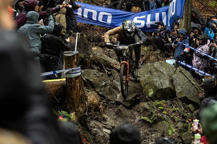 Mountain biker navigating a rocky forest course with spectators and banners in the background.