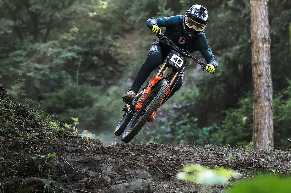 A mountain biker in full gear, including a helmet, goggles, and a dark teal jersey, riding an orange and black full-suspension bike over a dirt jump in a forest setting.